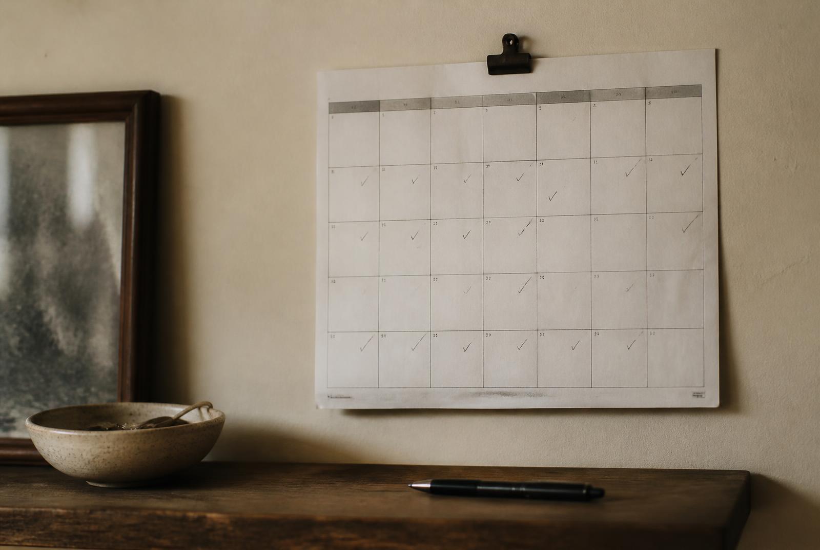 Wall calendar with small tick marks on completed days and a pen resting nearby, tracking a new habit without perfectionism