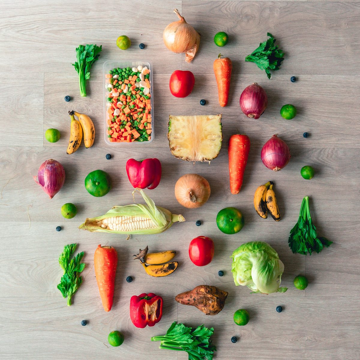 Colourful fresh fruit and vegetables — carrots, onions, tomatoes, peppers, bananas, limes, corn, cabbage and blueberries — laid out on a wooden surface